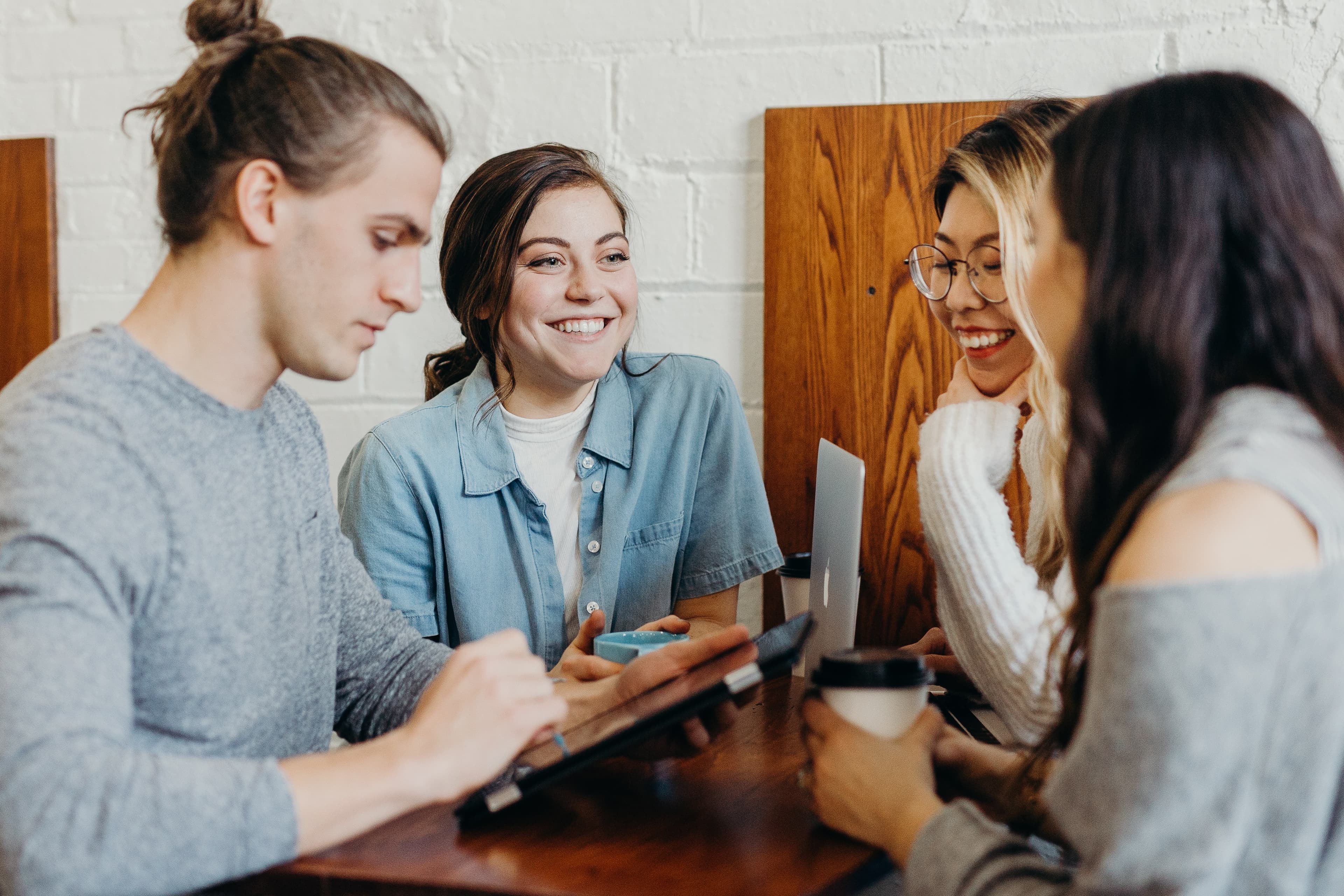 Colegas de trabalho conversando e sorrindo durante um intervalo. Há três mulheres e um homem, todos interagindo uns com os outros ou com dispositivos tecnológicos.